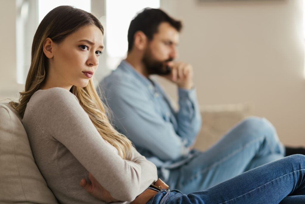 Worried young woman sitting on sofa at home and ignoring her boyfriend who is sitting next to her